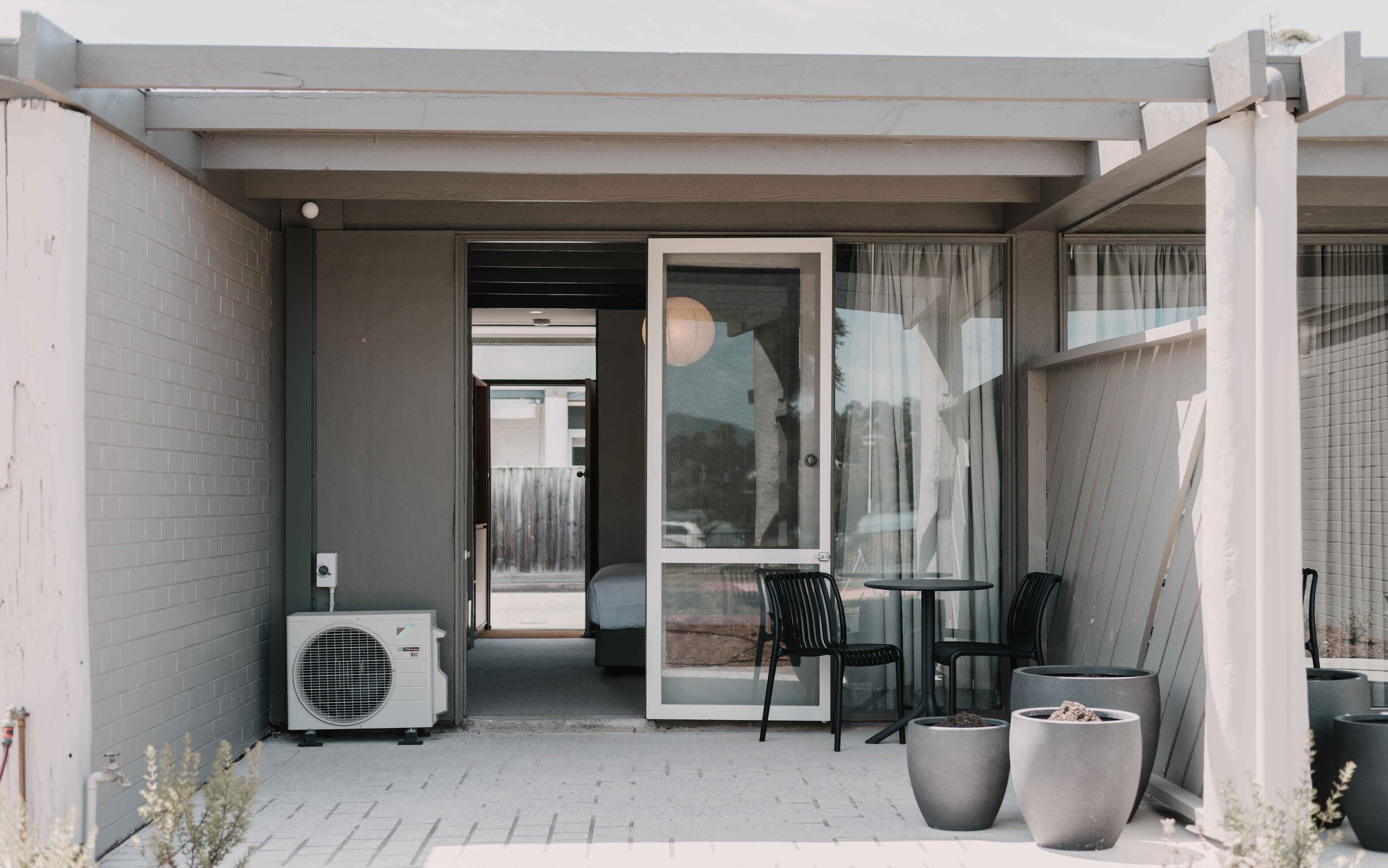A small outdoor seating area with a table and chairs, a potted plant, and a glass door leading to a room.