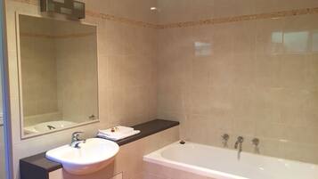 Un baño con lavamanos blanco, espejo, bañera y paredes con baldosas color crema.