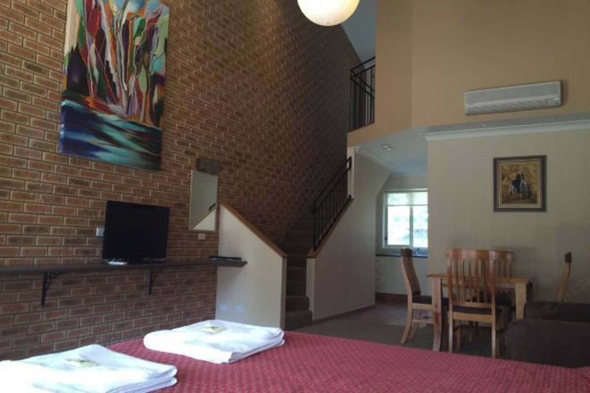 A hotel room with a brick wall, a staircase, a flat-screen TV, a bed with a red bedspread, and a painting.