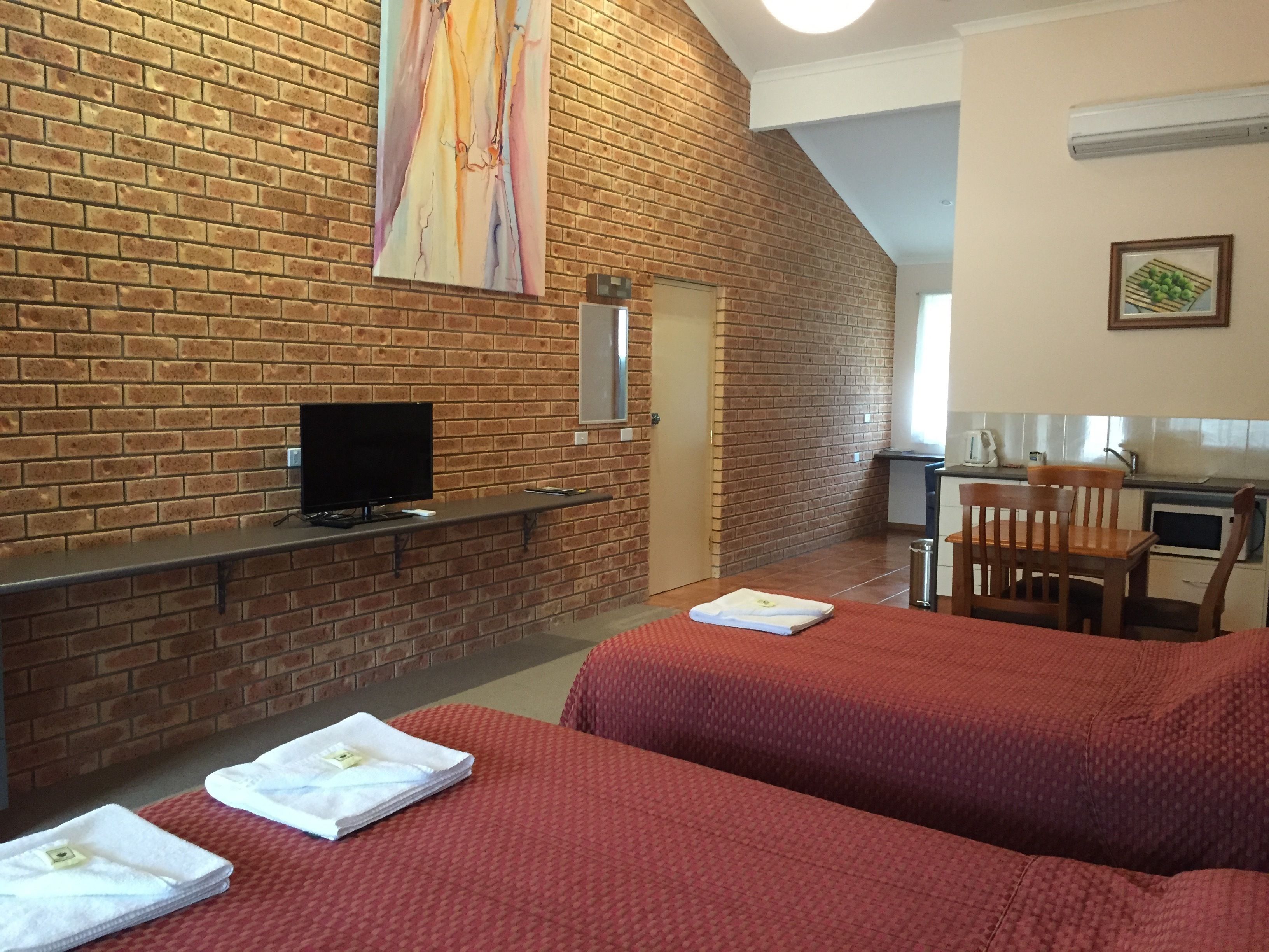 A room with a brick wall, a flat-screen TV, two beds with red bedding, a wooden chair, and a small kitchenette.