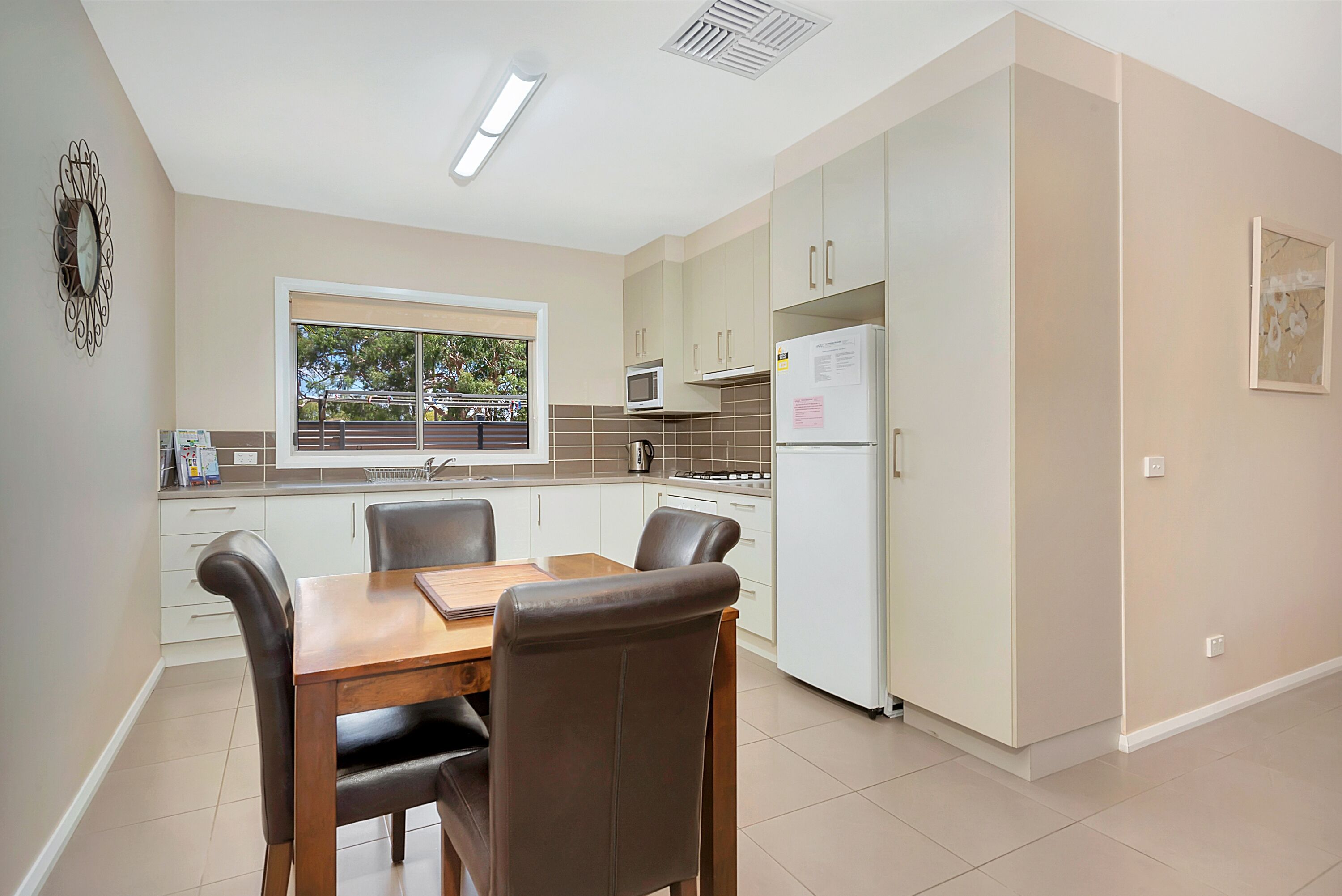 A modern kitchen with white cabinets, a dining table with chairs, a window with a view of greenery, and a refrigerator with a note on it.