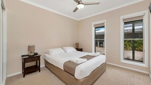 Un dormitorio con cama, dos mesitas de noche, ventilador de techo y ventana con vista a follaje.