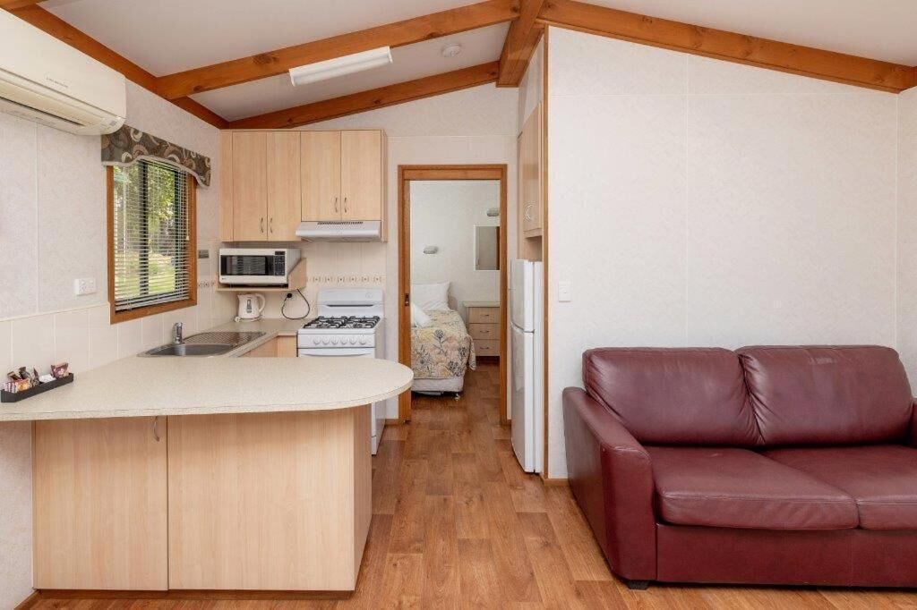 Une cuisine compacte avec des meubles en bois, un four à micro-ondes, une cuisinière et un réfrigérateur. On y trouve un petit îlot avec un évier, et dans l’espace salon, un canapé en cuir rouge.