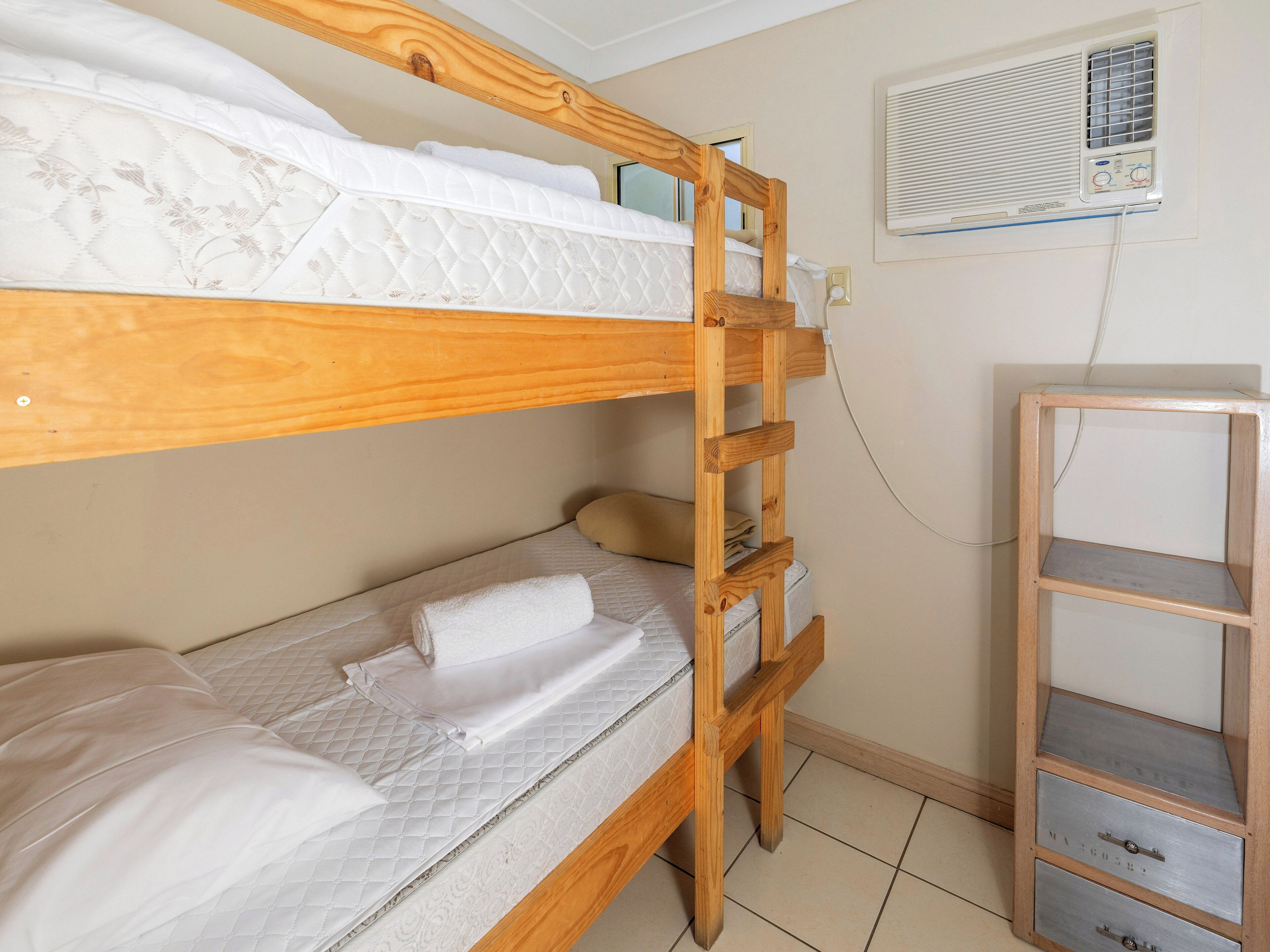 Ein Stockbett mit Holzleiter, ein kleiner Nachttisch und eine Wand-Klimaanlage.
