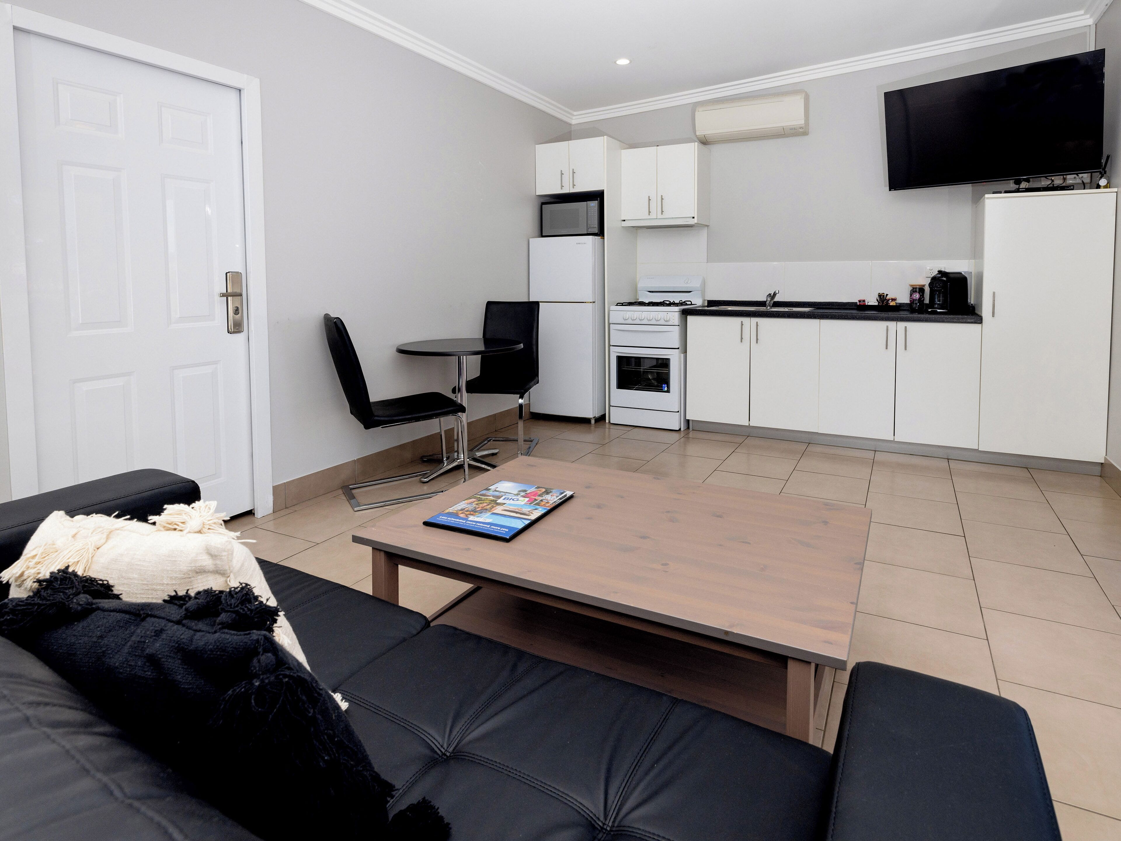 A compact living space with a sofa, a coffee table, a dining area with chairs, a kitchen with cabinets and appliances, and a TV mounted on the wall.