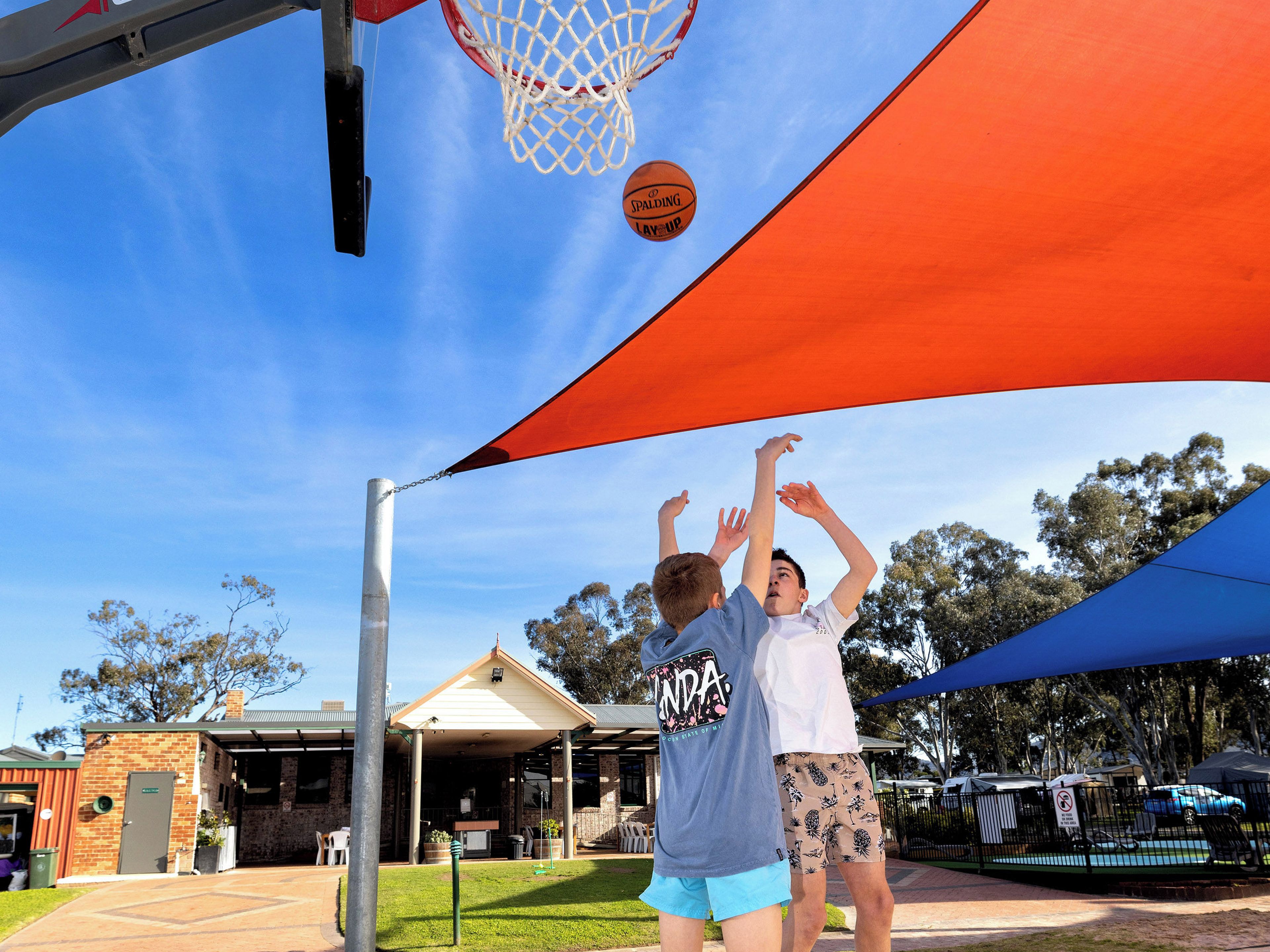 Dos personas jugando baloncesto bajo una zona con sombra y un edificio al fondo.