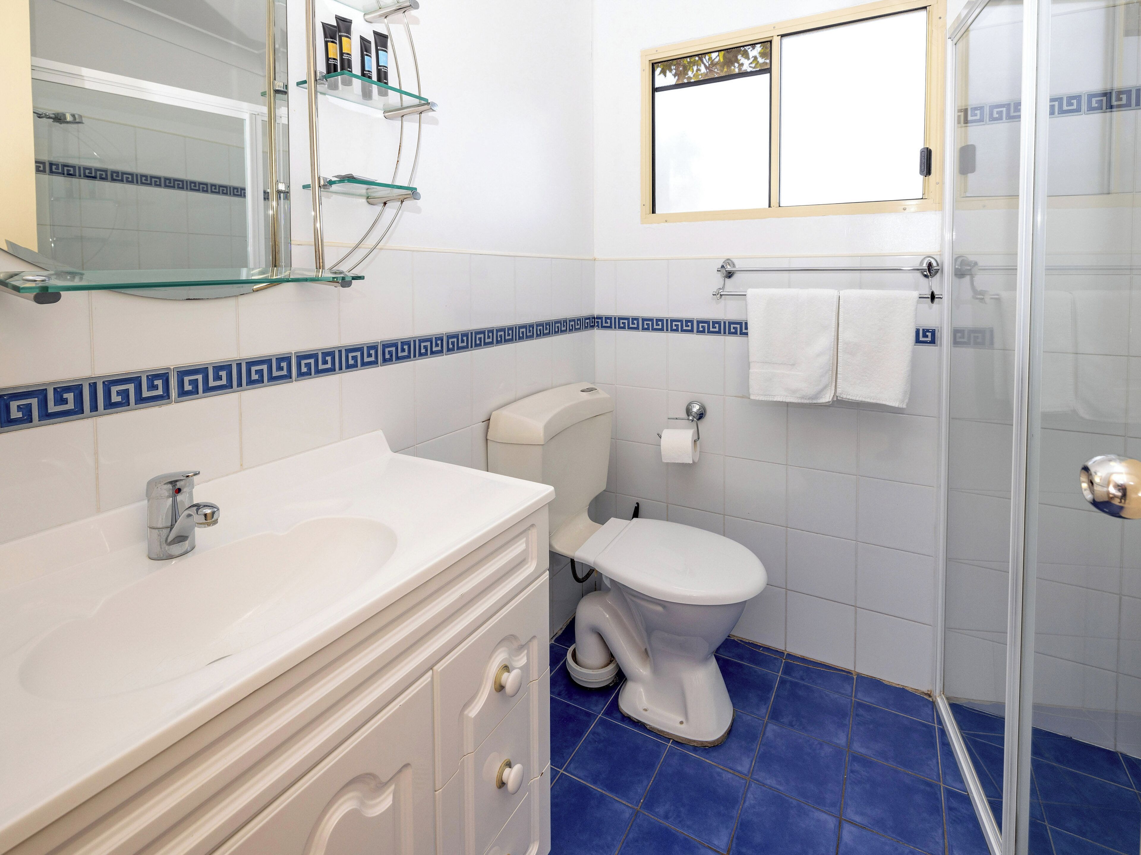 Un baño con un lavabo blanco, un inodoro, cabina de ducha con mampara de vidrio y un espejo con repisas superiores.