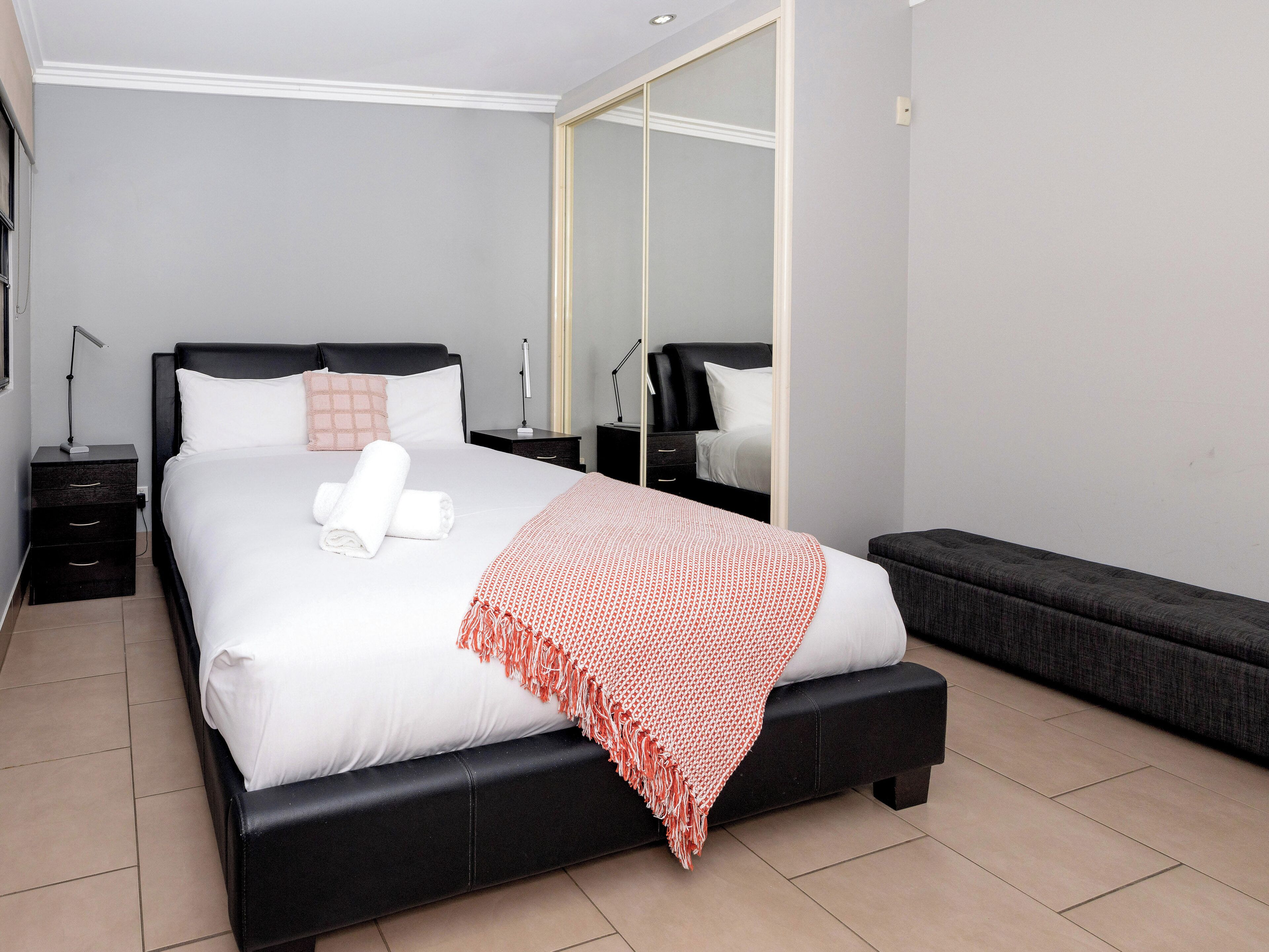 A neatly made bed with white linens and a red and white patterned throw blanket, a black bench, and a large mirror on the wall.