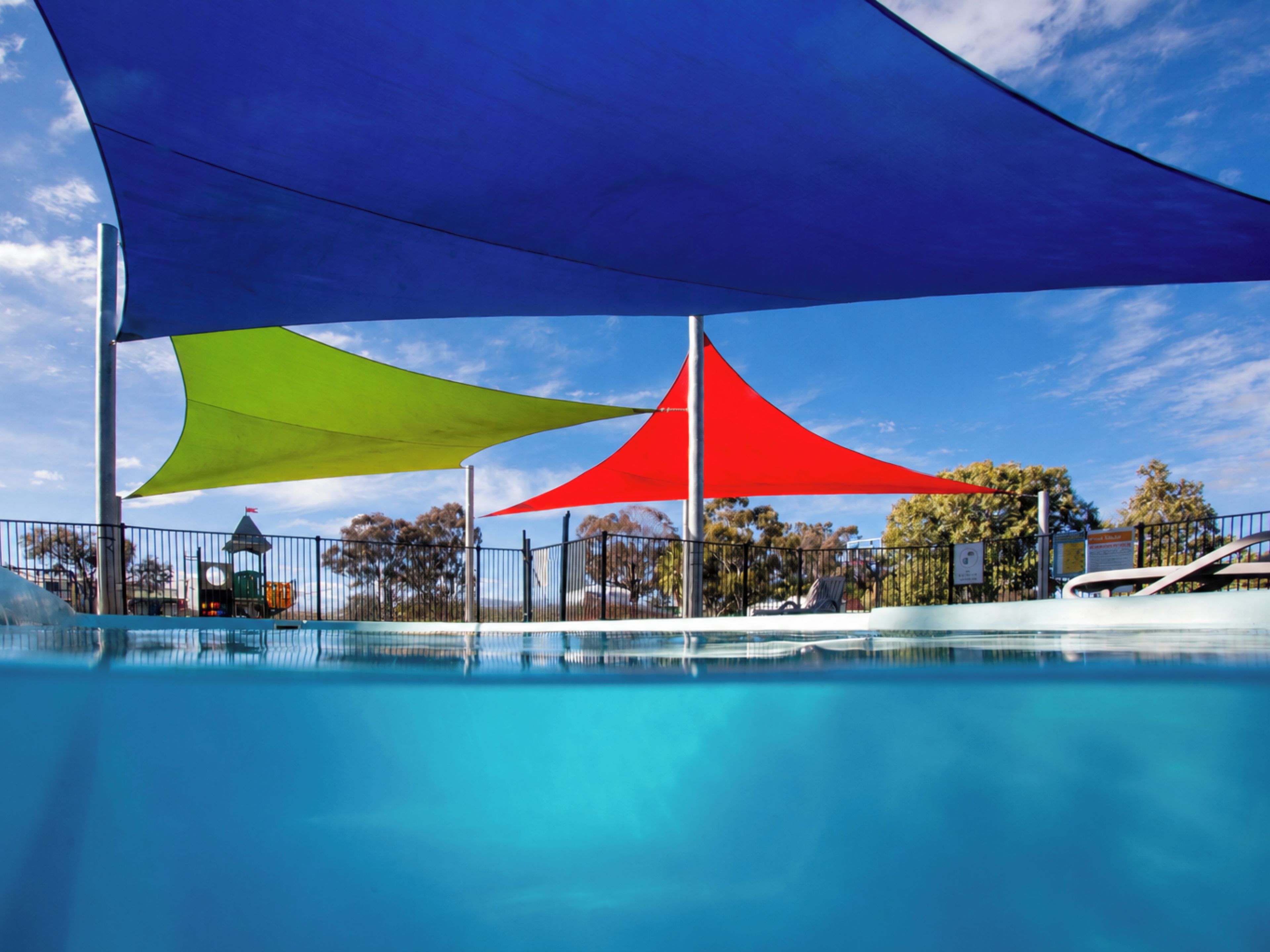 A swimming pool with a clear blue water surface, surrounded by a fence and shaded by colorful fabric canopies.
