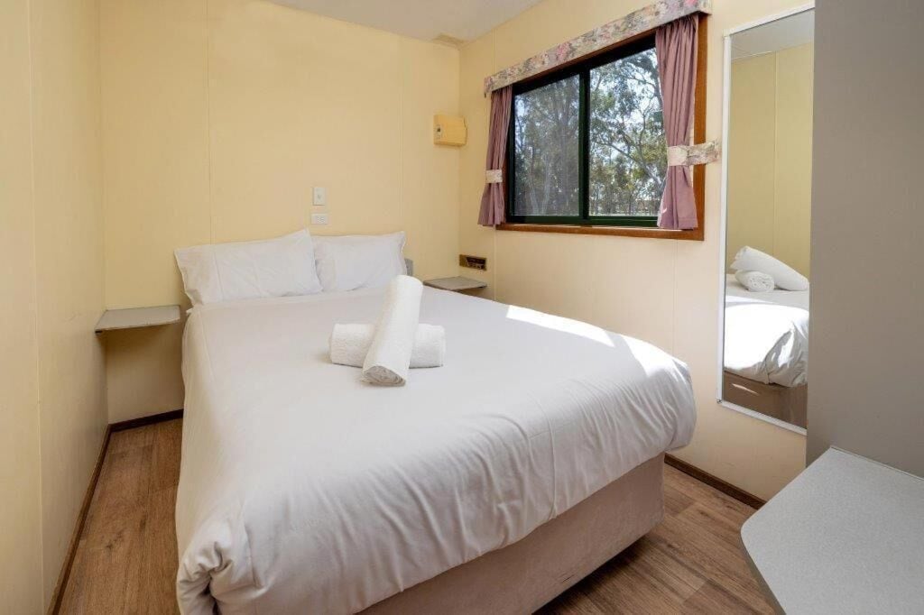 A neatly made bed with white linens and rolled towels, a small bedside table, and a window with curtains.