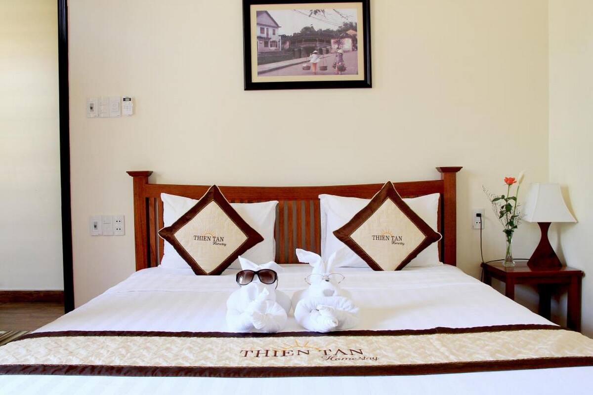 Habitación de hotel con cama, dos almohadas con el logotipo de 'THEN TAN Homestay', un cuadro enmarcado en la pared, mesita de noche con lámpara y un jarrón con flores.