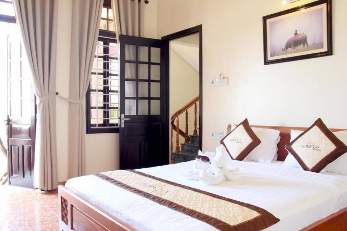 Habitación de hotel con una cama grande, cabecera de madera, un cuadro enmarcado en la pared y vistas al exterior a través de una ventana con cortinas.