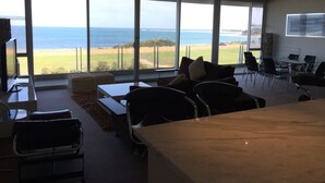 A spacious room with large windows offering a view of the sea and a golf course. There is a seating area with a table and chairs, and a television on a stand. - Onshore Torquay (Torquay)