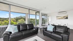 A modern living room with two black leather sofas, a white coffee table, and large windows offering a view of greenery. - Onshore Torquay (Torquay)