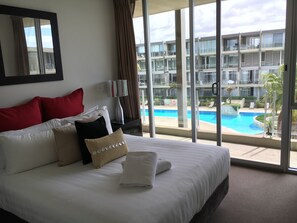 Een hotelkamer met een groot bed, een nachtkastje, een lamp en uitzicht op een zwembad en een gebouw.