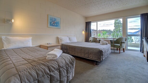 A hotel room with two beds, a small table, a chair, and a window with a view of greenery. - Lakeside Motel - Queenstown (Queenstown)