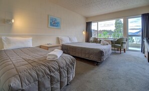 A hotel room with two beds, a small table, a chair, and a window with a view of greenery. - Lakeside Motel - Queenstown (Queenstown)