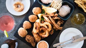 Diversas preparaciones de mariscos fritos y aperitivos sobre una tabla de pizarra negra, acompañados de bebidas y utensilios.