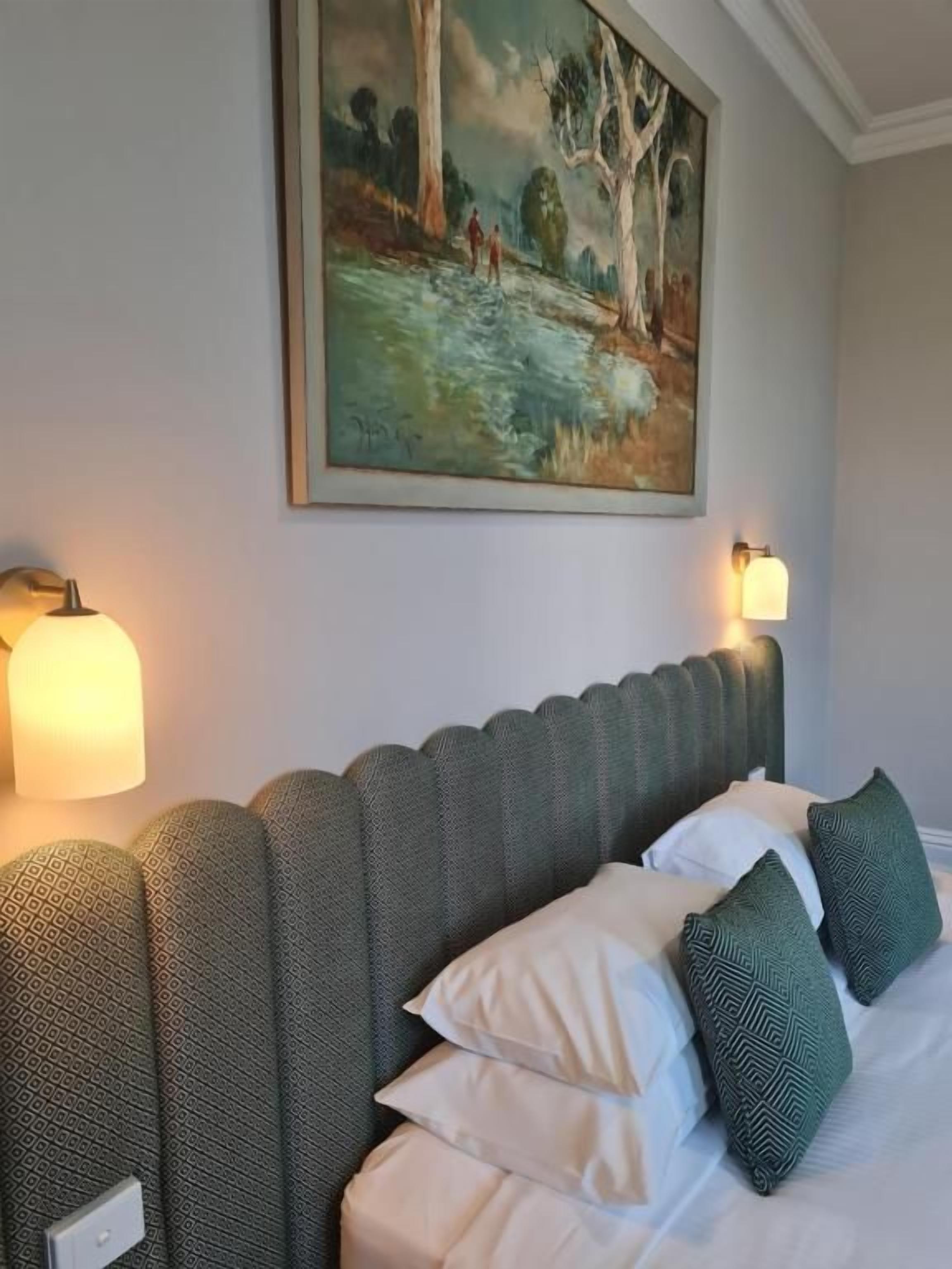 Een opgemaakt bed met patroonkussens, een hoofdbord met een getande rand, en een schilderij erboven.