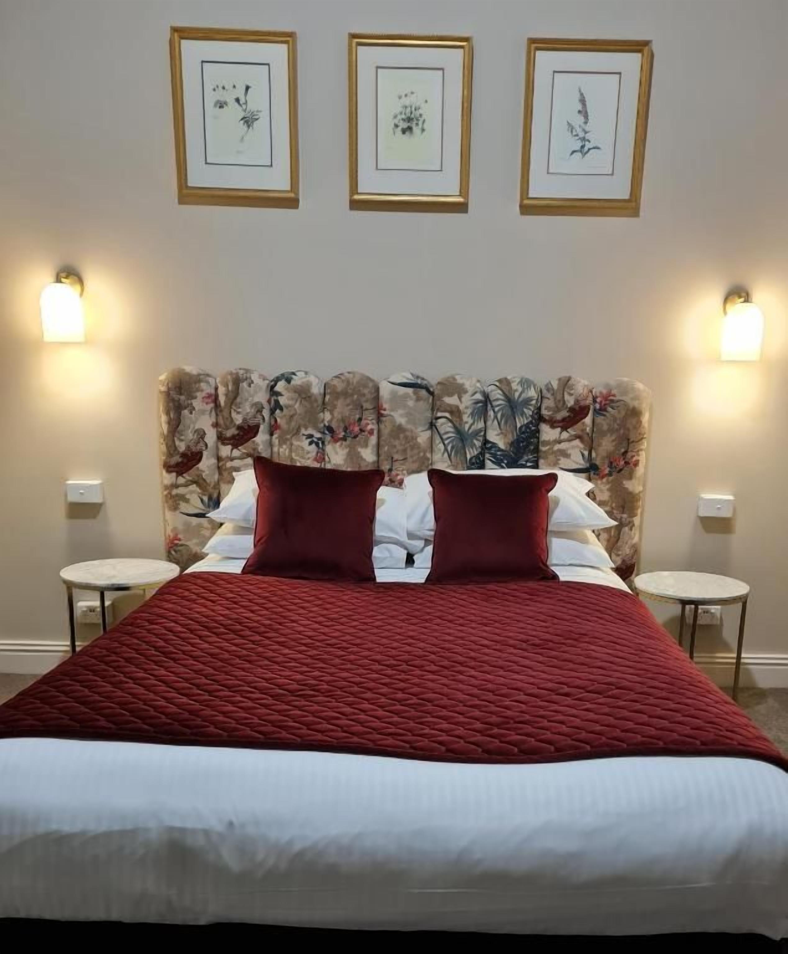 Une chambre d’hôtel avec un grand lit, une tête de lit à motifs floraux, deux tables de chevet et des lampes fixées au mur.
