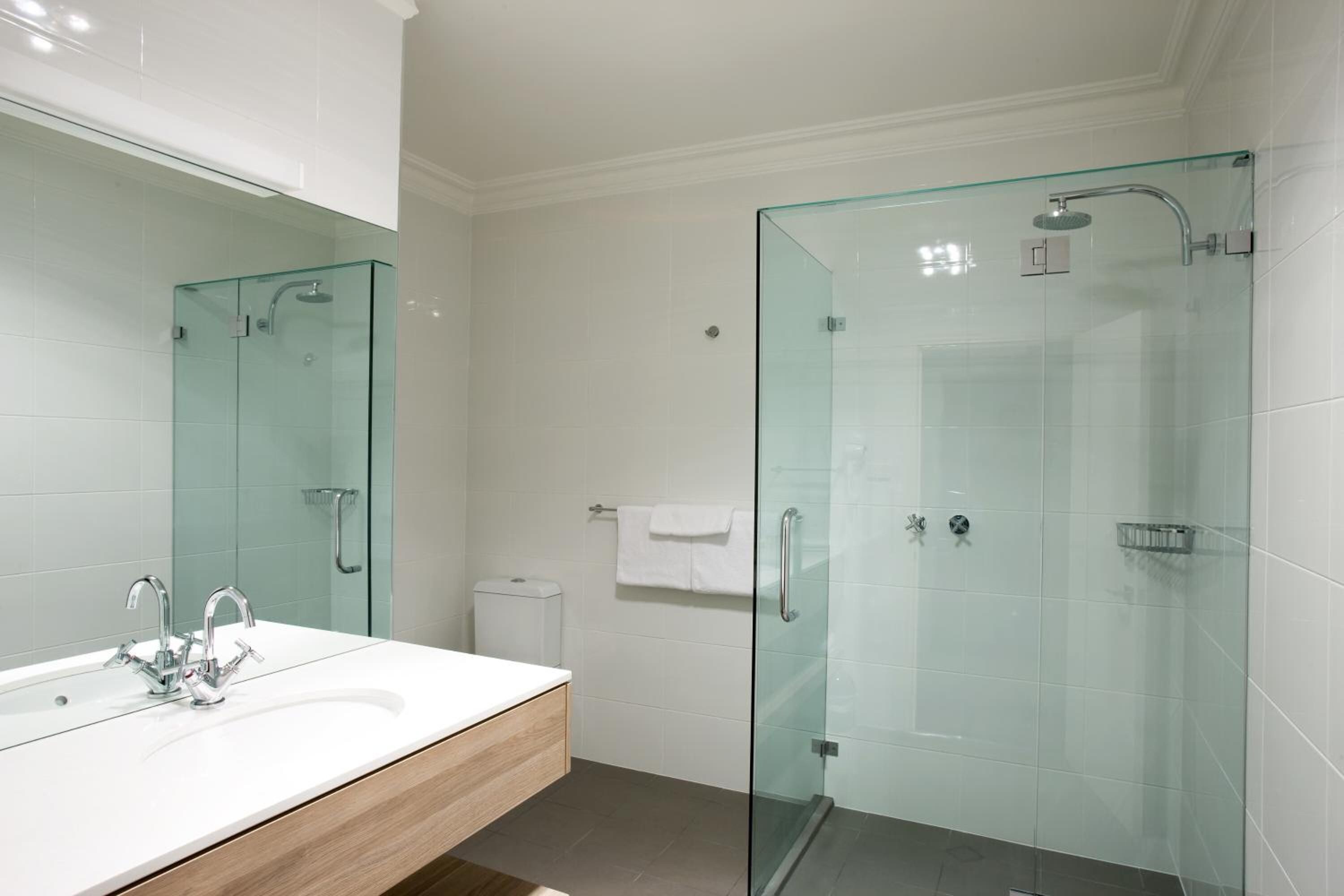 Un baño moderno con ducha envuelta en vidrio, lavamanos blanco con grifería cromada y tocador de madera.