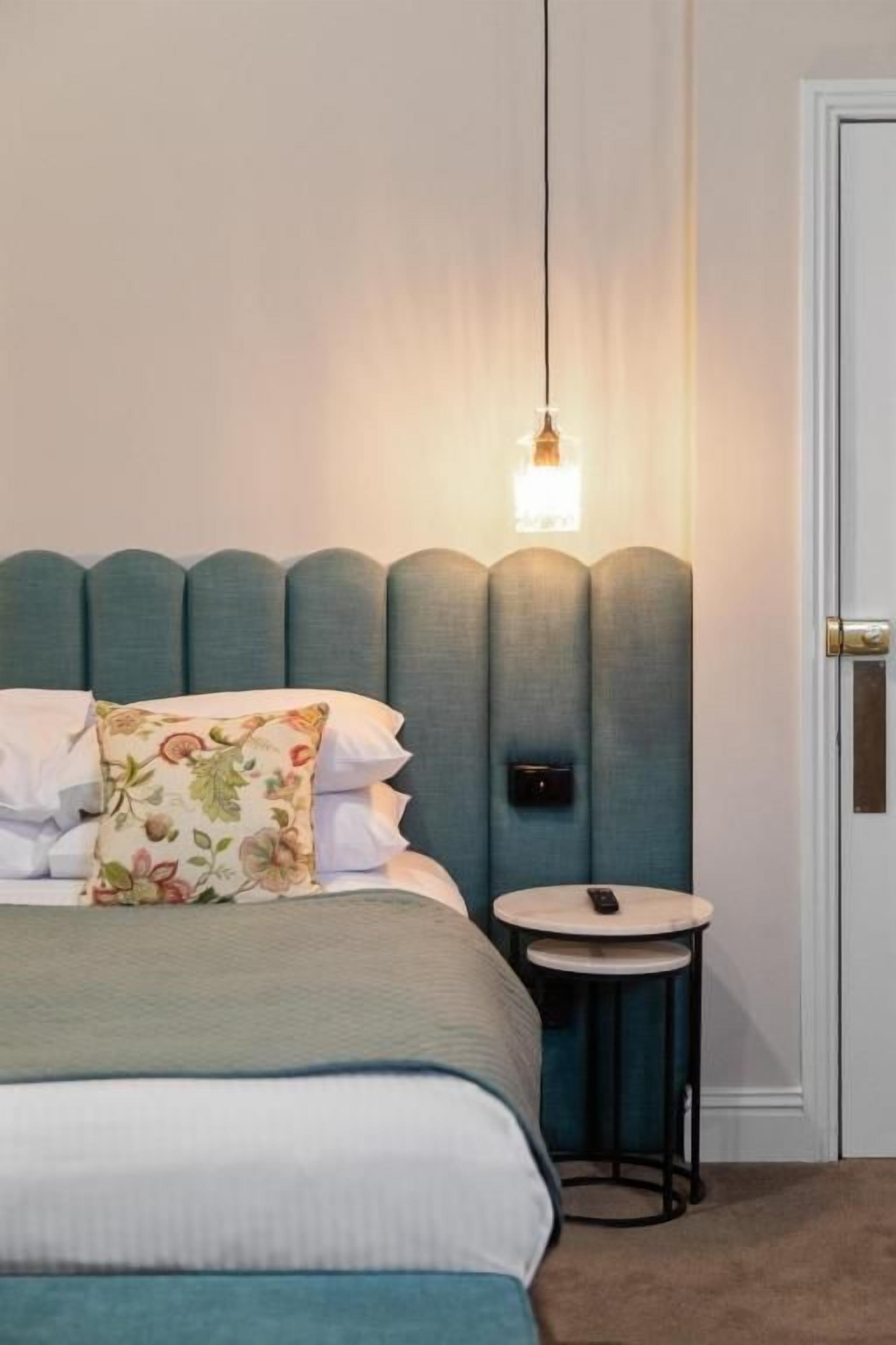Een net opgemaakt bed met een bloemenkussen, een nachtkastje met een afstandsbediening, en een hangende lamp.