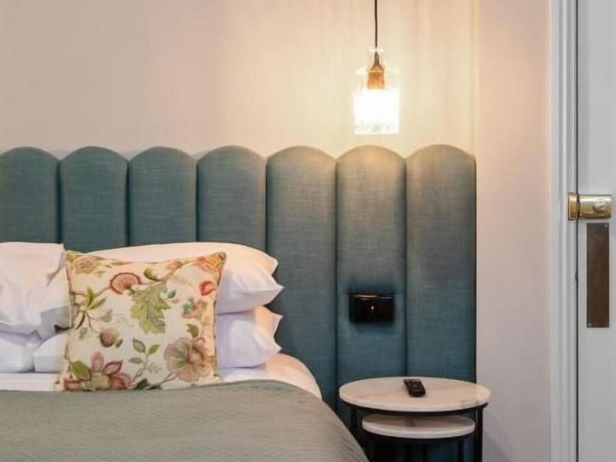 Een net opgemaakt bed met een bloemenkussen, een nachtkastje met een afstandsbediening, en een hangende lamp.