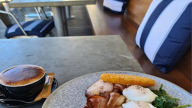 A breakfast plate with eggs, bacon, mushrooms, spinach, and a croquette, accompanied by a cup of coffee on a table in a cafe setting.