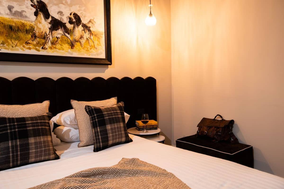 A neatly made bed with plaid pillows, a framed painting above it, a bedside table with a glass of wine and a book, and a handbag on a nightstand.