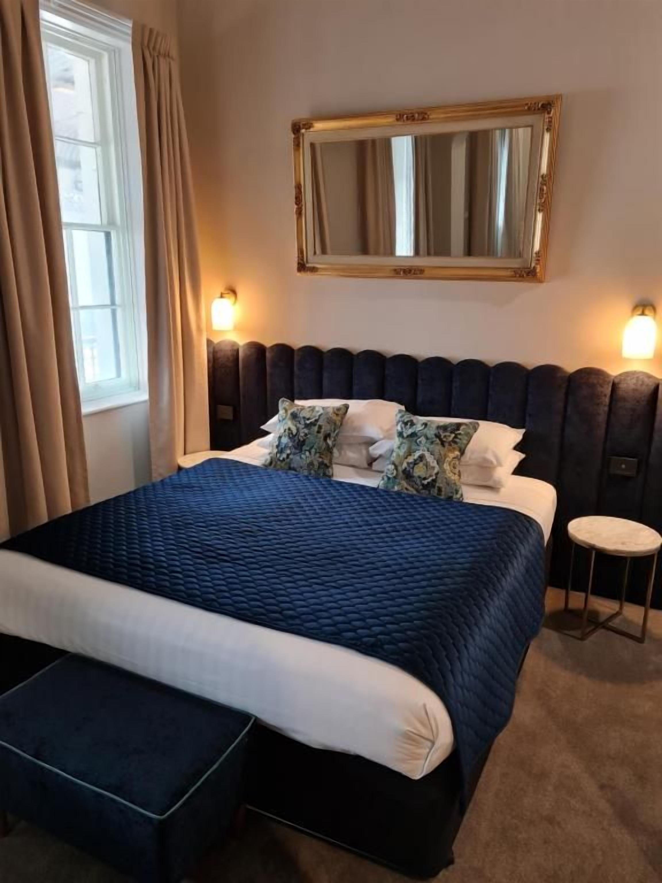 Habitación de hotel con una cama grande, una mesita redonda, un espejo sobre la cama y una ventana con cortinas.