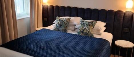 Habitación de hotel con una cama grande, una mesita redonda, un espejo sobre la cama y una ventana con cortinas.