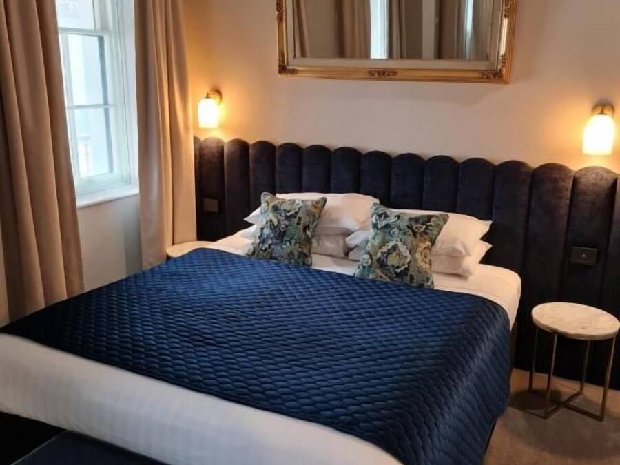 Habitación de hotel con una cama grande, una mesita redonda, un espejo sobre la cama y una ventana con cortinas.