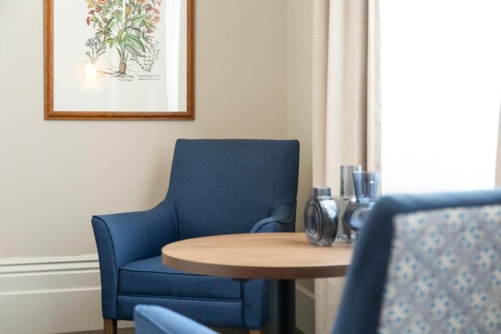 Una stanza con una poltrona blu, un tavolo rotondo in legno e una stampa botanica incorniciata appesa al muro.