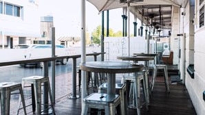 An outdoor seating area with tables and stools, a covered section with a view of the street, and a building with a 'bank' sign in the background.