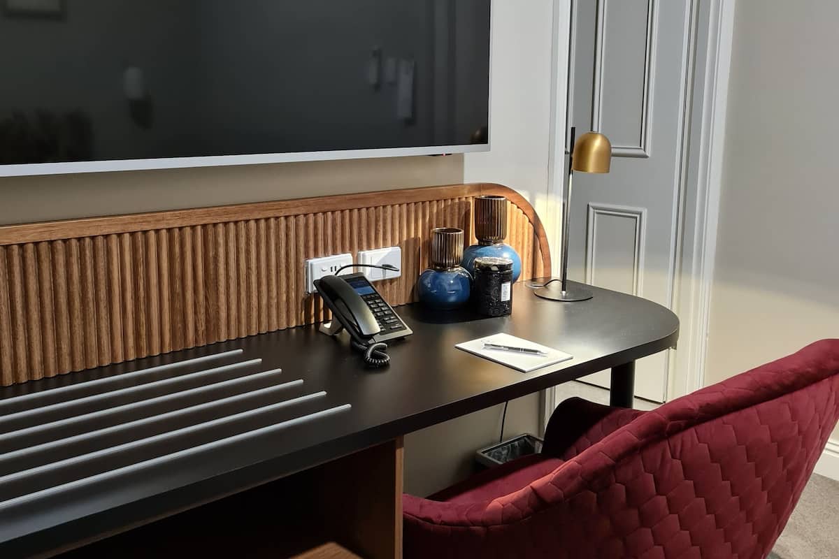 A room with a large flat-screen TV mounted on the wall, a wooden desk with a telephone, a red cushioned chair, and a door with a gold handle.