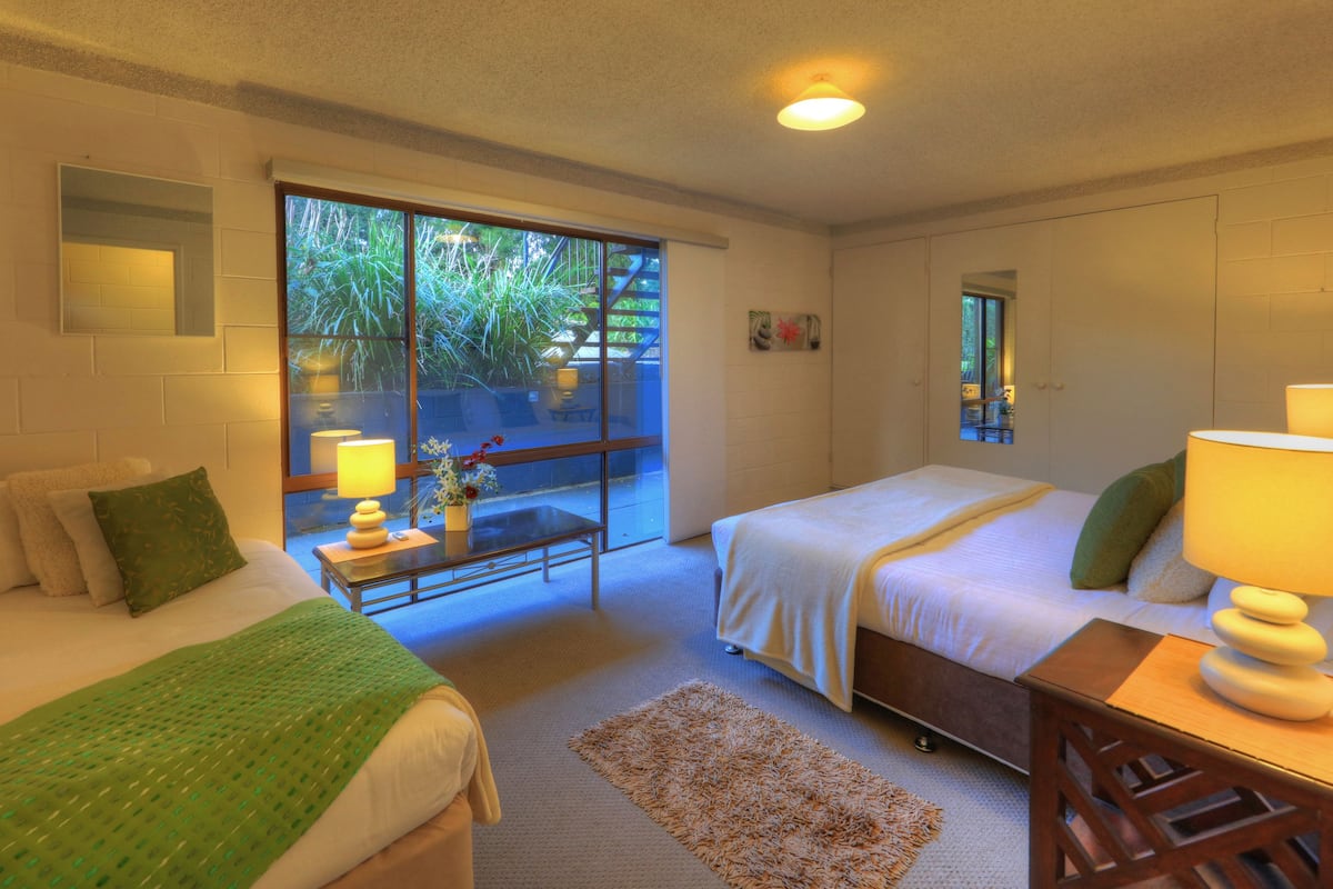 A hotel room with two beds, a small table, a chair, a window with a view of greenery, and a ceiling light.