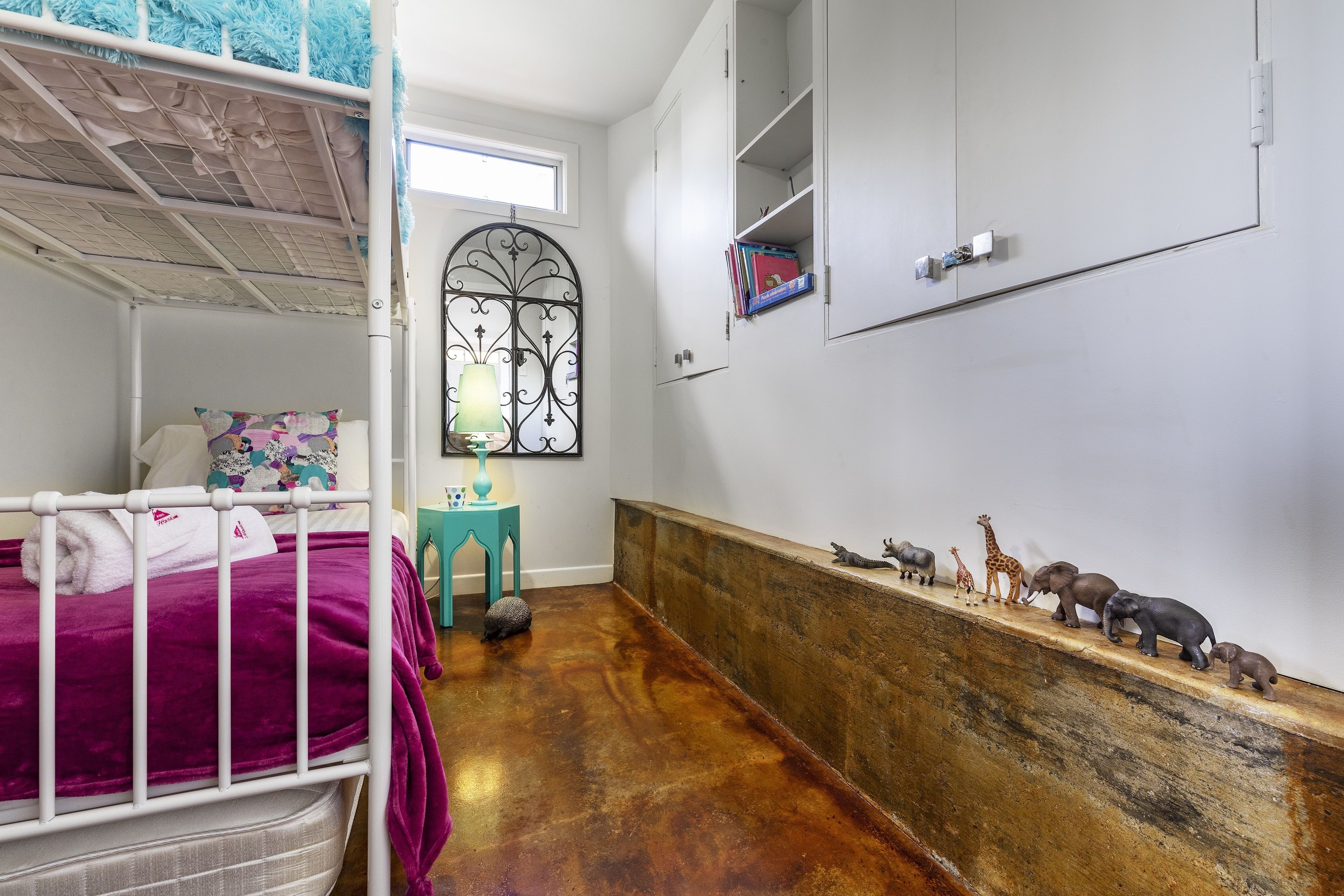 A bedroom with a bunk bed, a small table with a lamp, a shelf with animal figurines, and a window with a decorative iron grille.