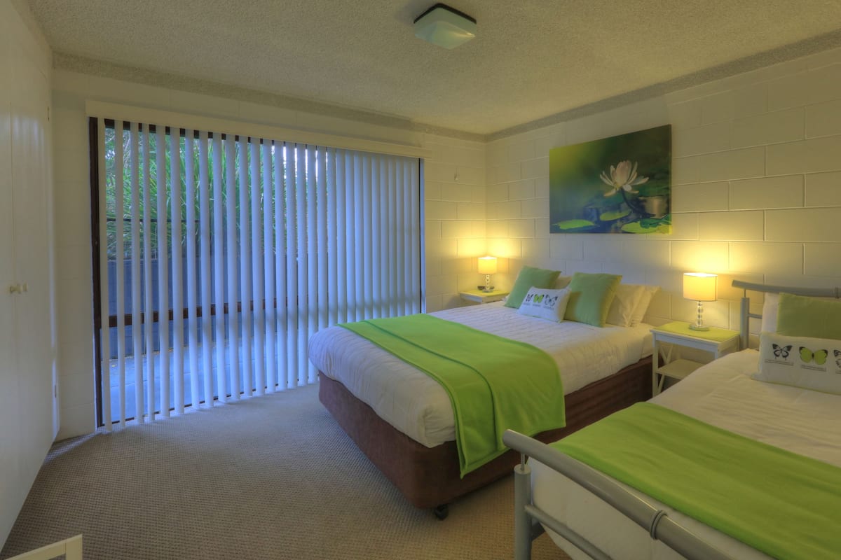A hotel room with two beds, a painting on the wall, and a window with blinds.