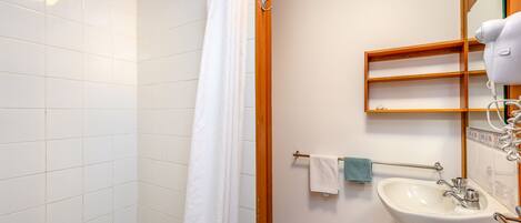 A bathroom with white tiled walls, a wooden door, a white sink with a mirror, and a hairdryer on the wall.