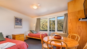 A room with a bed, a small table with chairs, a window with a view of a forest, and a painting on the wall. - Kasees Apartments & Mountain Lodge (Thredbo)
