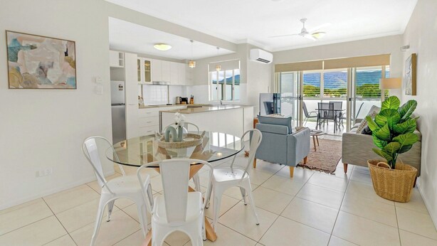 A modern kitchen with white cabinets, a dining area with a glass table and white chairs, and a living area with a sofa and a large plant.