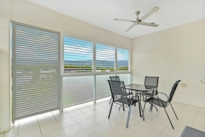 A balcony with a table and chairs, a ceiling fan, and large windows with blinds. - Trinity Beach Retreat (TRINITY BEACH)