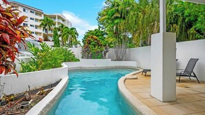 A swimming pool with a curved edge, surrounded by lush greenery and a white building in the background.