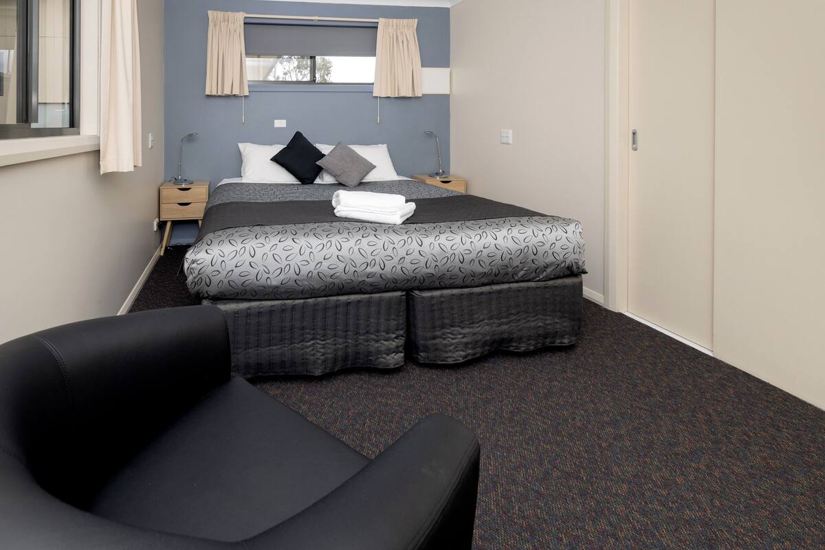A hotel room with a bed, a chair, a nightstand, and a window with curtains.