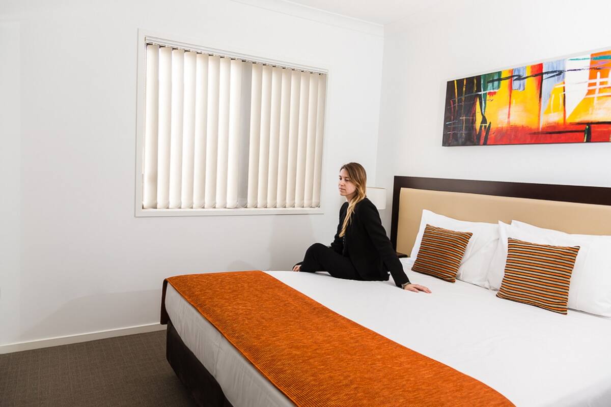 Une chambre d’hôtel avec un lit, une fenêtre avec des stores, un tableau au mur et une moquette marron.