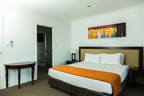 A hotel room with a bed, a nightstand, a table, and a painting on the wall. - Gladstone Heights Executive Apartments (Gladstone)