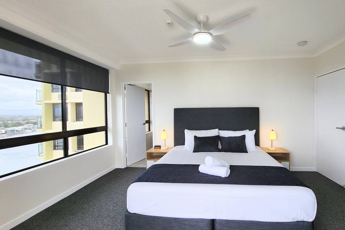 Habitación de hotel con una cama grande, ventilador de techo, mesita de noche con lámpara, una puerta y una ventana con vistas a edificios.