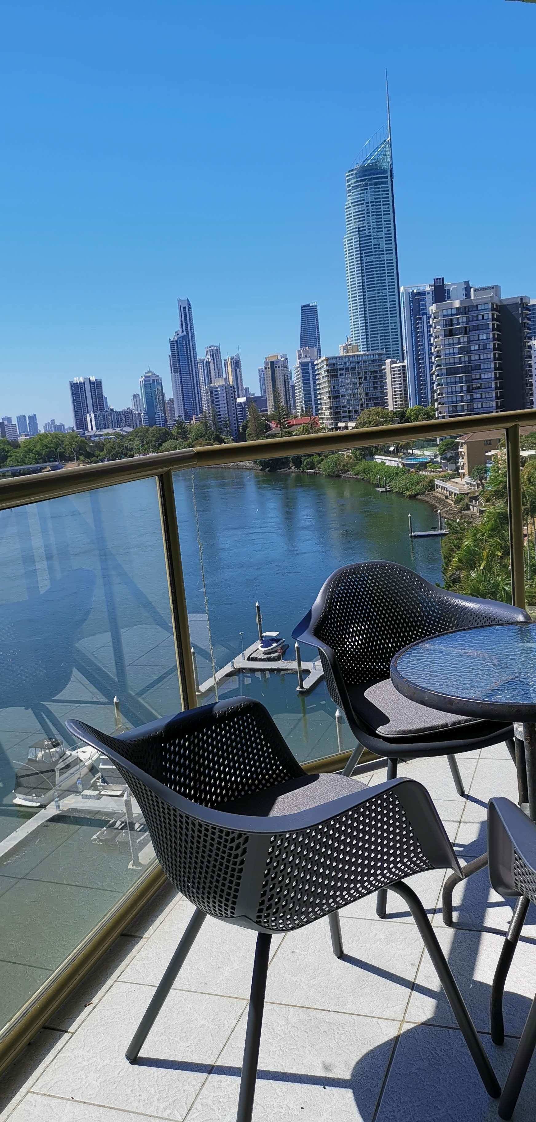 A balcony with two chairs and a table overlooking a river with boats, set against a city skyline.