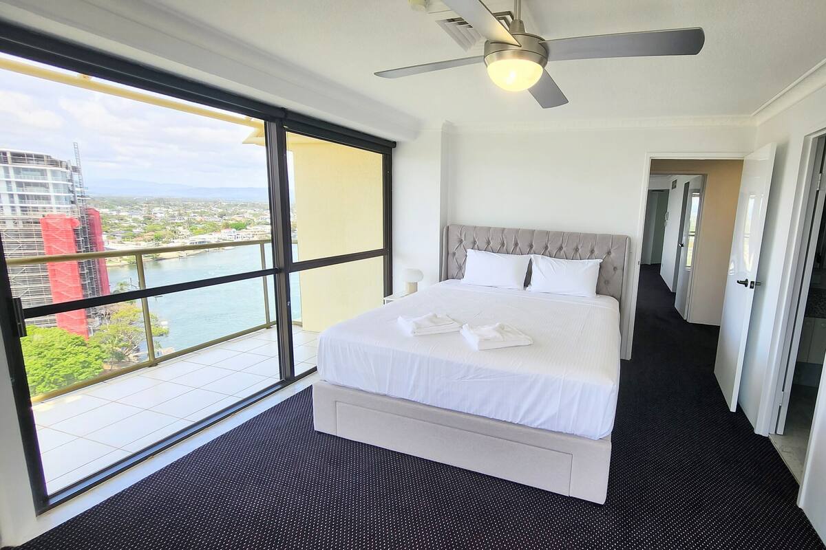 Habitación de hotel con una cama grande, un ventilador de techo y vistas de la ciudad a través de un amplio ventanal.