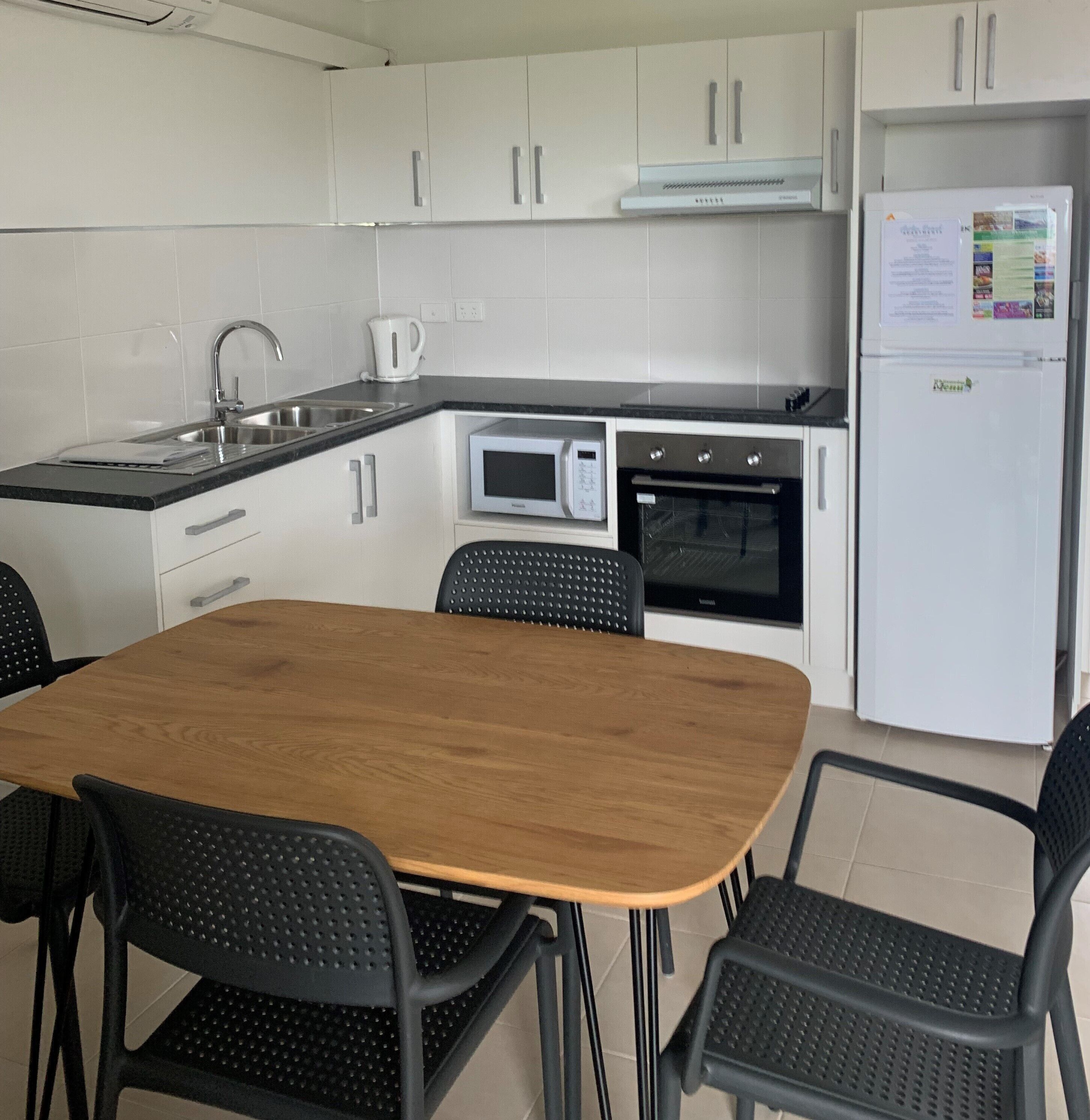 Een kleine keuken met witte kastjes, een magnetron, een oven en een koelkast. Er staat een houten tafel met vier stoelen.